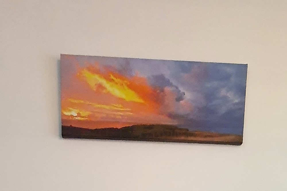 A framed sunset photograph on the treatment-room wall above the massage table, with warm orange light on the left and dark, stormy cloud on the right